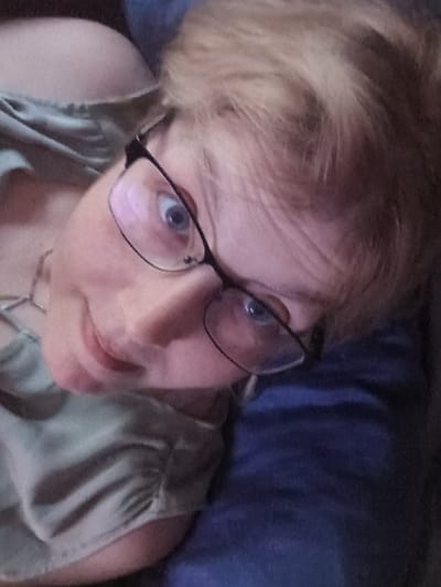 An upward-angle portrait depicts an adult individual with blonde hair and a fair complexion. The subject wears black-framed eyeglasses, through which the right eye, appearing blue and wide, is prominently visible. A slight smile is present on the lips. The individual is dressed in a light olive-green top featuring a gathered or ruched shoulder detail. A silver-colored chain necklace is partially visible around the neck. To the right and lower right of the subject, a dark blue fabric, possibly upholstery or bedding, forms an indistinct background element. The scene appears to be indoors. No specific identifying features are present within the image to pinpoint a precise location within the city of Libramont, Belgium.