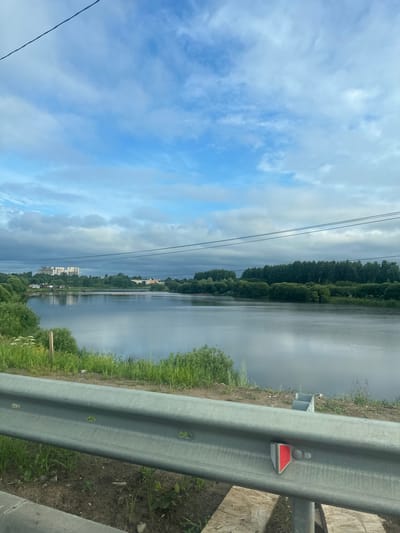 The image depicts a calm river scene with a cloudy sky. In the background, on the left side, are several tall buildings that appear to be apartment complexes. A dense line of trees borders the opposite bank of the river. In the foreground, a metal guardrail runs along a dirt embankment covered in green grass and some small plants. The guardrail has several rectangular cutouts and a red reflector. The overall impression is a serene, possibly suburban or rural landscape on a partly cloudy day. This view is consistent with locations near the town of Bratovshchina, Russia, known for its natural beauty and proximity to water bodies.