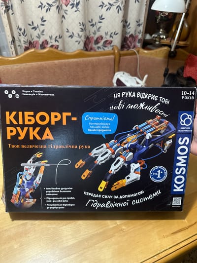 The image shows the front of a boxed toy kit named "KIBORG-RU KA" in Ukrainian. The box features an illustration of a robotic hand and highlights that it is a hydraulic hand. Text on the box indicates that it is a science, technology, engineering, and mathematics kit for ages 10-14. The background appears to be a wooden surface, possibly a table or shelf. The overall scene is indoors, with part of a curtain visible in the upper portion of the image. There are no people or specific urban environmental elements visible that would indicate a location in Nova Dofinivka, Ukraine.