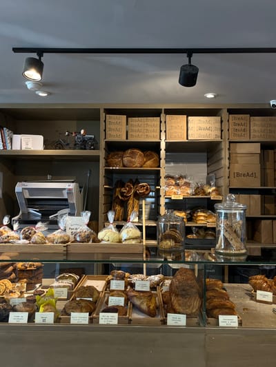 The image depicts a bakery in Ciudad Santa Catarina, Mexico, located at the coordinates 25.658 latitude, -100.439 longitude. The bakery is well-lit with overhead track lighting and features a glass display case with shelves stacked high with pastries. There are a variety of breads and pastries on display including croissants, breads, and other baked goods, arranged neatly in rows. Some of the items are labeled with prices and descriptions in Spanish. A silver commercial slicer can be seen behind the counter, suggesting baking operations are taking place on-site. The bakery's walls are lined with shelves displaying more baked goods, including numerous packages of bread, a few of which are labeled as "Bread." The walls are painted a light beige, providing a neutral backdrop for the brightly lit and vibrant display of bakery goods.