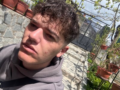 A selfie photograph shows a person with curly brown hair and fair skin, wearing a gray hooded sweatshirt. The background reveals an outdoor space with potted plants along a wall constructed of irregular stones. A metal structure supporting vines is also visible. The location appears to be a garden or patio setting, but a more specific determination of its location in Delchevo, Bulgaria is not possible from this image alone.