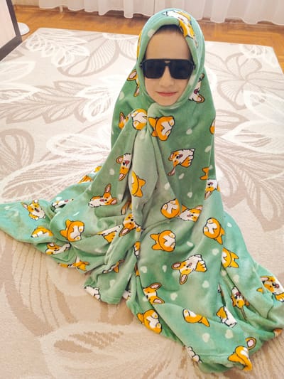 A child, wearing a light green blanket with a corgi pattern and large black sunglasses, sits cross-legged on a light-colored carpet. The blanket is draped over their head and body, obscuring most of their form. The carpet has a subtle floral pattern. In the background, a wooden floor is visible, partially covered by sheer white curtains. The scene appears to be indoors. There is no indication of the location being in Biaroza, Belarus.