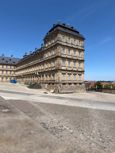 The image depicts the New Residence in Bamberg, Germany, under a clear blue sky. The grand Baroque palace, characterized by its extensive stone facade, multiple stories, and elaborate roofline with dormer windows and decorative elements, occupies a significant portion of the frame. In front of the palace, a large, paved courtyard, made of cobblestones in various patterns and colors, stretches out. To the left of the palace, a blue portable building is visible. Two park benches are situated against the stone base of the palace. Beyond the palace, the rooftops of the city of Bamberg, with their characteristic red tiles, are visible, along with some trees and greenery. The scene is tranquil, with no people visible, suggesting a peaceful daytime ambiance.