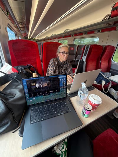 A woman is working on two laptops on a train. She is seated at a table in the train car, with red seats visible behind her. On the table are two laptops, a water bottle, and a cup with a lid. The train car appears to be modern, with overhead luggage compartments and bright lighting. The woman is wearing glasses and a leopard-print top. She is focused on her work, with her hands on the keyboard of one of the laptops. The scene is well-lit and the image is sharp, suggesting it was taken in daylight. The overall impression is one of productivity and travel. The location is a train traveling through the English countryside.
