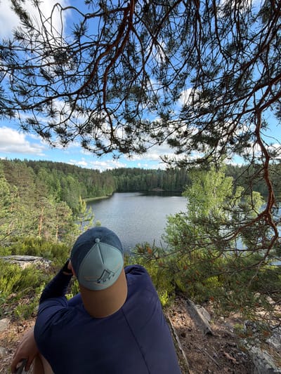 A person wearing a navy blue long-sleeved shirt and a baseball cap is seated on a rocky outcrop, looking out at a lake. The cap is teal and tan with an emblem. The lake is surrounded by a dense forest of pine and deciduous trees under a bright, cloudy sky. The image is taken from a high vantage point, looking down over the person and the landscape. The location is not identified as being in the city of Ruch'i, Russia.