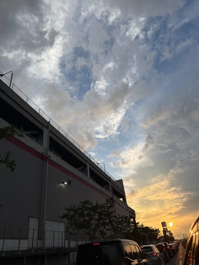 The image depicts an exterior scene at sunset in Sumiyoshi Minamimachi, Higashinada Ward, Kobe, Japan. The prominent structure on the left is the Costco Wholesale store, characterized by its multi-level concrete parking garage atop a large facility with corrugated grey metal siding and a distinct red horizontal stripe running along its upper facade. A single exterior light fixture is illuminated on the grey wall. Several small trees are visible at the building's base. The sky above features a mix of grey and white clouds with visible patches of blue, transitioning to a bright orange and yellow hue on the right horizon, indicative of a sunset. A long line of vehicles, primarily passenger cars, including a dark-colored van in the immediate foreground, is stationary or slowly moving in traffic along a road that recedes towards the setting sun on the right. Distant structures, including what appears to be a large sign or billboard, are silhouetted against the bright horizon. No individuals are explicitly visible.