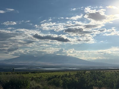 The image depicts a panoramic view of Mount Ararat in Armenia, seen from a distance. The foreground is dominated by lush green vegetation, possibly fields or orchards, with a variety of trees and bushes. In the middle ground, a body of water, likely a lake or river, stretches horizontally, reflecting the sky. Beyond the water, the majestic silhouette of Mount Ararat rises, its peak capped with snow. The mountain is partially obscured by clouds and dramatic rays of sunlight pierce through the cloudy sky, creating a dramatic and atmospheric scene. The sky itself is a dynamic canvas of blue, with scattered white and gray clouds, some illuminated by the sun, suggesting either sunrise or sunset. The overall impression is one of natural beauty and grandeur, with a sense of vastness and tranquility. The location in Paruyr Sevak, Armenia, is known for its scenic landscapes, and this image captures a quintessential view of the region.