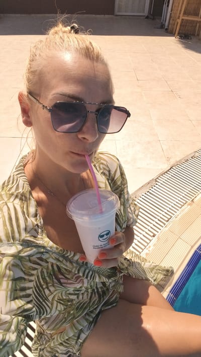 A woman with blonde hair pulled up in a messy bun and wearing oversized sunglasses is taking a sip of a light-colored beverage from a clear plastic cup with a pink straw. She is dressed in a tropical-print blouse. The woman is positioned near a swimming pool, indicated by the blue water and tiled edge visible in the lower right portion of the image. The background suggests an outdoor setting with tiled flooring and some bamboo-like structures, possibly in a resort or a backyard area. The lighting indicates it is daytime, likely sunny. The woman's hands are visible, with one holding the cup and the other resting near her lap. The overall scene depicts a leisurely moment, possibly at a vacation spot.