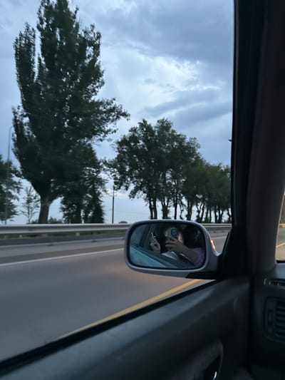 In a car moving on a highway, an individual is reflected in the side mirror. The person is holding a cigarette in their left hand and a smartphone in their right hand, looking at the phone. They are wearing a purple sweater. The background visible in the mirror shows a line of trees. Outside the car, the scene depicts a highway with a guardrail on the left side, and a line of trees and a cloudy sky. The image appears to have been taken from inside the car, looking out the passenger side window, with the side mirror in the foreground. The location is identified as Dostuk, Kyrgyzstan.