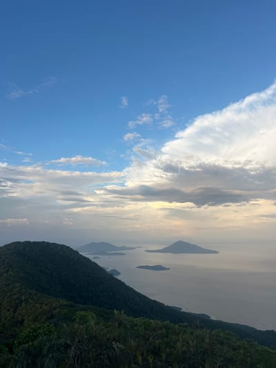 The image depicts a panoramic view from a forested mountain slope overlooking a body of water with several islands. The sky is partly cloudy with dramatic white clouds against a blue background. The foreground is dominated by dense green foliage of trees and plants characteristic of a tropical or subtropical mountainous region. The middle ground features the dark green slopes of the mountain extending down towards the water. In the distance, several islands of varying sizes rise from the calm, grayish-blue water. The overall scene conveys a sense of natural beauty and tranquility. There are no people or man-made structures visible in the image. The location is El Cacao, El Salvador, a region known for its lush landscapes and coastal views.