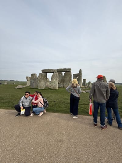The image displays Stonehenge, an ancient monumental stone circle, situated on a green plain near Amesbury, Wiltshire, United Kingdom, under an overcast sky. A paved pathway occupies the foreground. On the left side of this pathway, an adult male in a grey jacket, dark pants, and sunglasses, holding a yellow brochure, crouches alongside an adult female in a pink jacket and light blue jeans, who carries a black backpack. A child in a red jacket is positioned between them, embracing the female. This group appears to be posing for a photograph. To the right, several individuals stand on the pathway, their backs mostly towards the viewer, observing the monument. One female wears a light-colored bucket hat, a grey top, dark wide-leg pants, and leopard-print shoes. Adjacent to her, a male in an orange baseball cap, a grey shirt, and blue jeans holds an orange shopping bag. A thin, dark rope barrier separates the paved path from the expansive green lawn surrounding Stonehenge. The large megalithic stones of the monument, some with horizontal lintels, are centrally featured in the midground against the flat, open landscape.