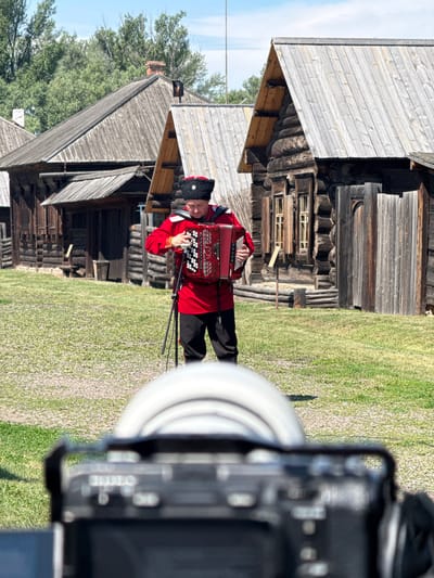In the city of Shushenskoye, Russia, a man dressed in a traditional red Cossack outfit with a black fur hat is playing an accordion. He is standing in front of several old wooden log houses with weathered wooden roofs. The houses appear to be part of a historical village or museum. The ground is covered in green grass and gravel. A camera lens is visible in the foreground, out of focus, suggesting the scene is being filmed or photographed. The sky is clear and blue with scattered clouds, and trees are visible in the background. The man is holding the accordion with both hands and appears to be actively playing it.