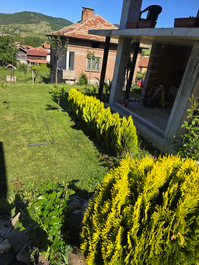 The image shows a landscape in Dorkovo, Bulgaria. In the foreground, there is a well-manicured lawn with two rows of yellow-green shrubs. In the background, there is a house under construction with a brick facade and a red tiled roof. A man is seen working on top of the house. Other buildings, including a smaller house with a red roof, are visible in the distance, and a green, hilly landscape rises in the background. A black, cylindrical object with a hose is lying on the lawn.
