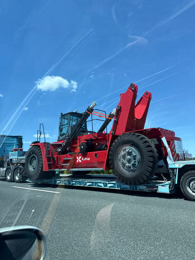 A large, red Kalmar container handler is on a flatbed truck. The handler has large tires, a driver's cab, and an articulating arm. The flatbed truck is on an asphalt road with a white line. The sky is blue with contrails and a few small clouds. The location is on the road, near Rotglà i Corberà, Spain.