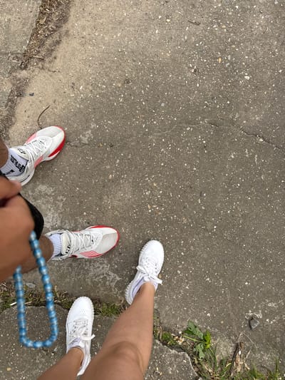 Two pairs of feet are positioned on a concrete surface. The concrete shows wear and tear with cracks and a small patch of green growth near the top right. One person is wearing white and red athletic shoes with white socks, holding a string of blue beads. Another person is wearing white shoes. The location appears to be Glojene, Bulgaria.