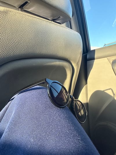 A pair of black-framed sunglasses with dark lenses rests horizontally on the upper leg of an individual, who is wearing navy blue fabric trousers. The scene is set inside a vehicle, with a light tan or beige upholstered seat back and headrest visible in the background, along with a matching door panel. Strong sunlight streams into the vehicle from the upper right, casting defined shadows on the interior surfaces. A section of clear blue sky is visible through the vehicle's window in the upper right quadrant. The specific location within Georgi-Dobrevo, Bulgaria, is not discernible from the provided image.