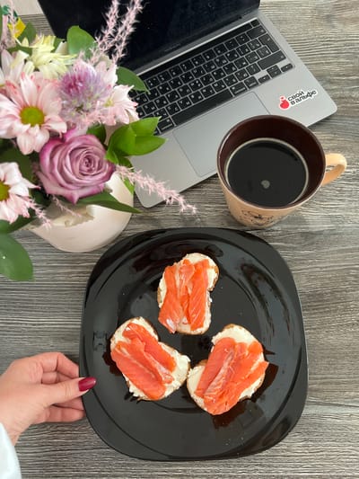 A high-angle medium shot captures a close-up of a table setting with food, a laptop, and flowers. In the foreground, a black plate holds three open-faced sandwiches topped with cream cheese and slices of smoked salmon. A woman's left hand, with dark red nail polish, is positioned near the edge of the plate, suggesting she might be reaching for a sandwich. To the upper right, a tan coffee mug filled with black coffee sits on a wooden surface. Adjacent to the mug is a silver laptop, partially open, with a sticker on its palm rest displaying a red heart with the Russian text "СВОИ В АЛЬФЕ." A bouquet of pink and purple flowers, including roses and chrysanthemums, is placed behind the laptop and to the left. The overall scene suggests a casual work-from-home or study session, possibly in a home environment in the village of Sovkhozny, Russia.