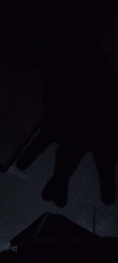 The image, taken at night in Ilogbo Eremi, Nigeria, is dominated by darkness. A silhouette of a building with a peaked roof is visible in the lower part of the frame. Above the building, darker shapes are discernable; a potential silhouette of a hand is the subject of the image. A tower is positioned toward the right side of the image. The sky is barely visible, appearing as a dark, grainy background behind the silhouettes. The scene lacks illumination, with only minimal light possibly from distant city lights.