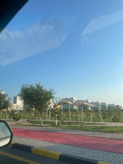 The image depicts a view of a modern park in the city of 30 Years of Kazakhstan, Kazakhstan, as seen from a vehicle. In the foreground, a red jogging path runs alongside a gray and yellow brick sidewalk. The park itself features lush green grass, scattered trees, and stylized white and wooden covered seating areas. Beyond the park, a line of tall, modern apartment buildings with light-colored facades stretches across the horizon under a clear blue sky. Reflections of clouds and possibly parts of the vehicle's interior are visible on the windshield.