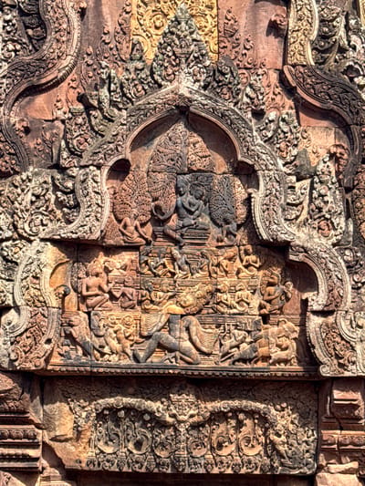 This image displays intricate stone carvings adorning the exterior of the Banteay Srei temple in Cambodia. The relief carvings depict various figures, mythological scenes, and decorative motifs. In the upper section, two seated figures, possibly deities or royalty, are centrally placed beneath an arched structure, surrounded by foliage and smaller seated figures. Below this, a larger tableau features numerous figures engaged in what appears to be a battle or procession, with some figures in dynamic poses and others kneeling or seated. The figures are rendered with detail in their attire and postures. The carvings are set within a richly ornamented architectural framework, characterized by scrolling patterns, floral designs, and layered elements that create a sense of depth and texture. The stone material exhibits a reddish-brown hue, indicative of sandstone, and shows signs of weathering and age. The overall scene is a testament to the detailed craftsmanship and artistic traditions of the Khmer Empire.
