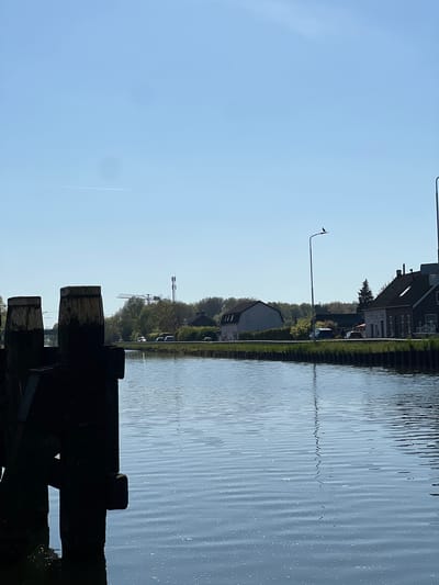 The image presents a tranquil daytime scene of a canal or river, likely located in Weert, Netherlands. The perspective is from the water's edge, looking across the waterway towards the opposite bank.
In the immediate foreground, a series of dark, weathered wooden posts or structures are silhouetted, partially obscuring the water beyond. The water itself, a calm expanse of light blue, occupies the central and left sections of the frame. It exhibits subtle ripples and reflections, catching the sunlight in shimmering patterns. A faint, thin, dark line, possibly a ripple or debris, extends horizontally across the water's surface.
The far bank, stretching across the middle ground, features a narrow strip of land populated by a mix of houses and dense greenery. Several residential buildings, characterized by dark roofs and light-colored facades, are nestled among abundant trees and bushes. A tall, slender street light stands out on the bank. Further in the distance, above the treeline, an industrial crane and a cellular tower are visible, suggesting a broader urban or semi-urban setting. A few parked cars can be faintly seen along the bank.
The sky, dominating the upper right portion of the image, is a clear, bright blue, indicating sunny weather. A distinct, straight white contrail from an airplane is visible high in the sky. The overall impression is one of a peaceful waterway environment under a clear sky, likely during the morning or afternoon. No people or explicit activities are visible, and there is no visible text.