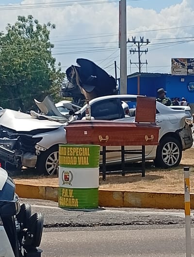 The image shows the aftermath of a severe car accident. A silver vehicle is totaled with extensive front-end damage, while another silver car has damage on the rear passenger side. A brown coffin rests on a stand in front of the intact car. A green and white drum with the words "ROAD ESPECIAL SEGURIDAD VIAL" is visible. A police motorcycle and a group of people are near the accident. The scene suggests a fatal collision.