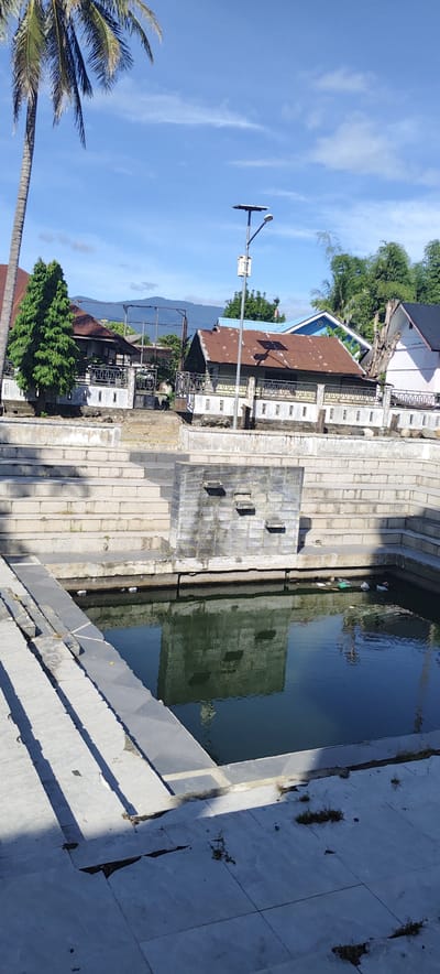 The image shows a terraced pool in Rheng, Indonesia. The pool is surrounded by concrete steps. A concrete structure with protrusions is in the middle of the pool. The water is still, reflecting the structure. Litter is present in the pool. In the background, there are buildings with metal roofs and a palm tree. A mountain range is visible in the distance under a clear blue sky. A utility pole stands near the buildings. The foreground features cracked concrete paving.