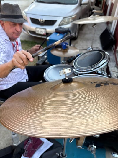 An older man in a grey fedora and striped shirt is seated outdoors, actively playing a drum kit. He holds two worn wooden drumsticks over the drums. In the foreground, a large bronze Paiste cymbal with visible concentric rings and a mounting screw with a wingnut and black felt pad is prominent. Behind the drum kit, a silver Toyota Corolla sedan with a Mexican license plate (PAR-1962 visible) is parked. A blue container and various drum components are visible in the mid-ground. To the far right, a light-colored wall with a brown top edge is present. A piece of red plaid fabric and a sheet of paper rest on a surface below the main cymbal. The scene appears to be a paved outdoor area, possibly a driveway, within the city of Chamilpa, Mexico.