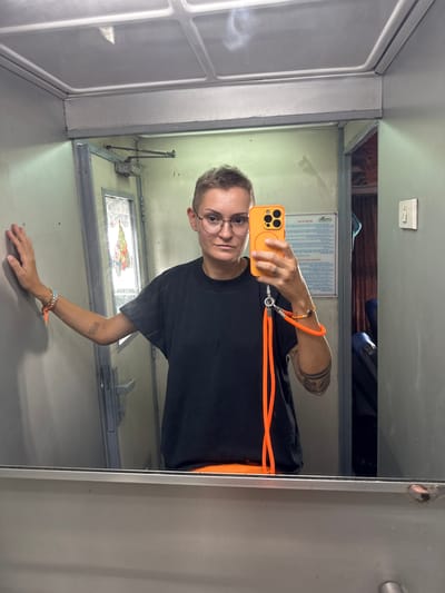 A person with short, buzzed hair, wearing glasses, a black t-shirt, and orange pants takes a selfie in a mirror. They are standing inside what appears to be a train compartment, with a door and a window visible in the reflection. The person's left arm is resting on a gray wall, and their right hand holds an orange smartphone. An orange lanyard is attached to the phone and drapes down. The environment is functional and utilitarian, typical of transportation interiors. The location is stated to be Vạn Giã, Vietnam.