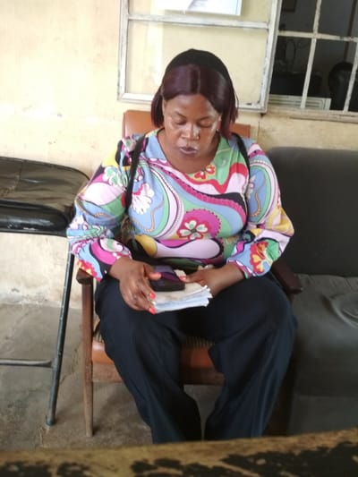 An African woman sits on a wooden chair in what appears to be an indoor waiting area. She is looking down at a stack of papers in her hands, which she is holding with her right hand. The woman has dark skin and dark hair that appears to be styled into a bob with bangs. She is wearing a colorful long-sleeved shirt with a psychedelic pattern, black pants, and her fingernails are painted red. A dark bag is slung over her left shoulder.
The background shows a pale yellow wall with a window that has metal bars. To the left of the woman is a black leather chair or bench, and to her right is a dark gray upholstered sofa. The floor is concrete. The scene suggests a government office, clinic, or other public building in Bisichi, Nigeria.