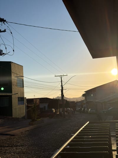 The image shows a street view in Pirenópolis, Brazil. The sun is high in the sky, casting long shadows. The street is made of cobblestones. There's a building to the left, a utility pole with many wires, and buildings on the right side of the street. Cars are parked along the street in the distance. A metal railing is in the foreground.