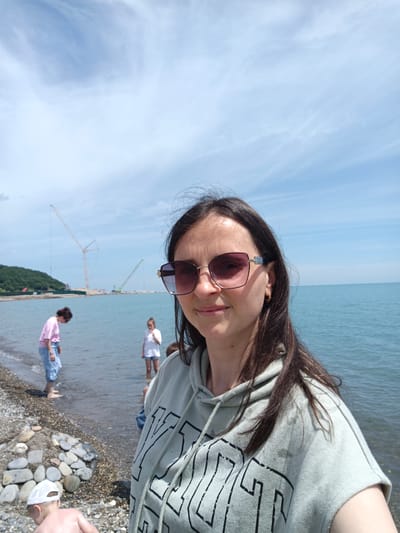 A female, likely in her 30s or 40s, with long dark hair and reflective sunglasses, is positioned in the foreground, facing and smiling slightly towards the camera. She wears a light green or grey sleeveless hooded sweatshirt with dark lettering. Her left ear displays a small golden hoop earring. The scene is a pebbly beach under a clear blue sky with sparse white cirrus clouds, indicative of sunny weather. Calm, blue-green water extends from the beach into the distance. In the left mid-ground, a woman with dark hair in a pink t-shirt and light blue jeans stands barefoot in the shallow water, looking downwards. Further right, a young girl in a white t-shirt and shorts stands in the water. In the bottom-left, a young child wearing a white baseball cap is partially visible on the pebbly shore, facing away from the camera. On the distant horizon, a green, tree-covered hill is present on the left, and multiple large construction cranes, including a tall yellow/orange lattice boom crane and a smaller green boom crane, are visible over the water, suggesting ongoing construction or port activity. A low barrier or breakwater extends from the land into the water near the cranes. The location is Krinitsa, Russia.