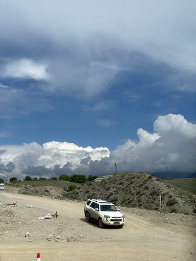 A white SUV is parked on a dirt road in a mountainous, rural setting. The car is positioned at an angle on the right side of the frame, with its front facing towards the left. The road is unpaved and appears to be made of gravel and dirt, with some larger rocks scattered along its surface.

The landscape is characterized by steep, rocky hillsides covered in sparse vegetation. A vast blue sky dominates the upper portion of the image, filled with dramatic, puffy white clouds, suggesting fair weather with potential for changes. The lighting indicates it is daytime, likely midday or early afternoon, given the brightness and the shadows.

There are several signs visible on the side of the road. One sign, partially obscured, shows a triangle with an exclamation mark, often indicating a warning. Below it, another circular sign with a red border displays the number "20", likely a speed limit. Another smaller sign with an "X" symbol is also present. Utility poles with wires run along the side of the road, indicating the presence of infrastructure.

In the distance, more hills and some scattered trees can be seen, with a hint of a valley or a wider expanse beyond. The overall impression is one of a remote, scenic location, possibly a popular route for off-road vehicles or hikers, given the road conditions and the presence of signs. No people are visible in the image, and the car appears to be stationary, suggesting it is either parked for a break or has stopped for a specific reason. The scene evokes a sense of adventure and the rugged beauty of the natural environment.