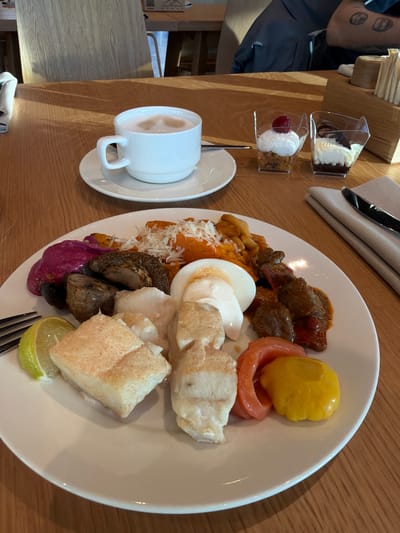 The image depicts a table setting with a plate of food, a cup of coffee, and two dessert cups. The plate contains a variety of items including what appears to be fish, meat stew, eggs, potatoes, mushrooms, and lime. A coffee cup sits on a saucer next to the plate. The dessert cups hold layered sweets. There is a hat, napkin, and silverware to the upper right of the plate. The table is wooden. The location, Озёрное, Russia, is not discernible from the image itself. There are no people other than an arm that appears to belong to a person wearing a blue shirt.