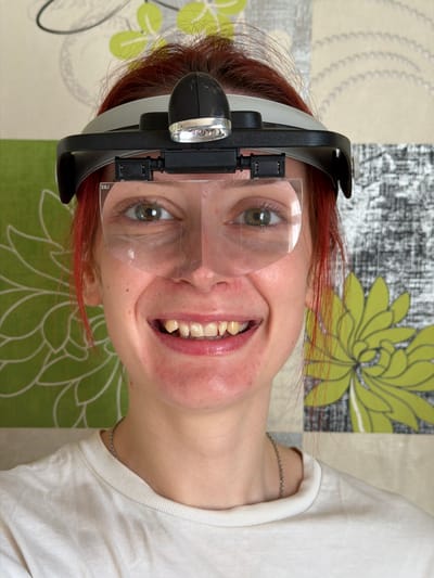 A woman with red hair and fair skin is smiling at the camera. She is wearing a white t-shirt and a headlamp with magnifying lenses. The background is a patchwork pattern of green and beige with stylized floral designs. She has freckles and a silver necklace. There is a silver bracelet on her left wrist. There is no information to suggest that the image was taken in Заболотово, Russia.