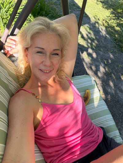 A woman with blonde hair and freckles is lying on a striped swing chair outdoors. She is wearing a pink tank top with decorative straps and dark shorts. A banana rests on the chair next to her. The background shows grass and gravel, suggesting a park or yard setting. The lighting indicates it is daytime with sunlight casting shadows.