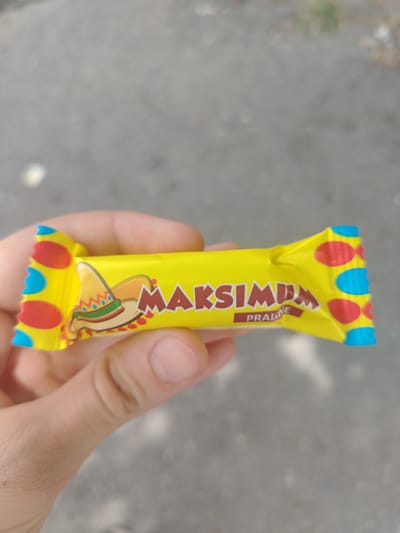 A person's hand is holding a yellow candy wrapper with "MAKSIMUM" written in brown letters. The wrapper has a colorful design with red, blue, and yellow circles and a depiction of a sombrero. The background is blurred asphalt. The image was taken in the city of Shaki, Azerbaijan.