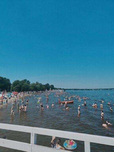 The image captures a wide, panoramic view of a crowded beach and lake in Nieporęt, Poland. The sky is a clear, bright blue. The foreground features a white wooden pier, with a portion of it visible at the bottom of the frame. Beyond the pier, the lake is filled with numerous people swimming, wading, and relaxing. The water is a deep blue, reflecting the sky.
The beach, visible on the left side of the image, is densely packed with sunbathers, umbrellas, and beach chairs. Lush green trees line the edge of the beach, forming a backdrop to the populated area. In the distance, across the lake, a shoreline with buildings and trees is visible, suggesting a populated area on the other side.
The people in the water are engaged in various activities: some are swimming, others are standing and socializing, and some are using inflatable toys. The scene is one of a bustling summer day at a popular recreational spot.