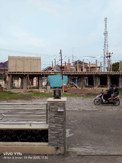 The image shows a partially constructed building in the city of Kecamatan Gabuswetan, Indonesia (latitude: -6.44, longitude: 108.028). The structure is made of concrete and wood and is in the early stages of construction, with the roof frame and some exterior walls in place. The ground is dirt and there is some construction debris scattered around. A blue tarp hangs from the partially built structure, with some wooden beams visible. In the foreground, a person is riding a pink motorcycle towards the camera. The environment is dusty and dry, with a clear blue sky in the background. A tall antenna is seen on the right side of the image. A metal gate is on the left side of the image, with a stone-covered pillar. The image appears to be taken from across the street.