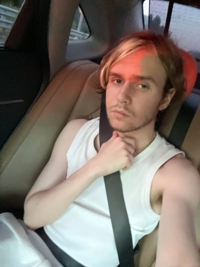 A young person with blond hair and light skin is seated in the back of a car. They are wearing a white tank top and a seatbelt. Their right hand is resting on their chin. A red light is reflecting on their forehead. The car's interior, including leather seats, is visible. Outside the car window, trees and a grey sky can be seen, suggesting an overcast day. The specific location of Bol'shoe Sareevo, Russia, cannot be determined from the visual information in the image.