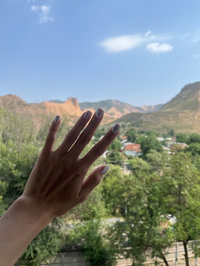 A hand with light-colored nail polish is held up in the foreground, obscuring part of a distant mountainous landscape and a small village. The sky is a clear blue with a few wispy white clouds. The village appears nestled in a valley with green trees and houses with red roofs visible. The mountains in the background are rugged and dry, with shades of brown and green. The overall scene suggests a sunny day with a view of a rural area.