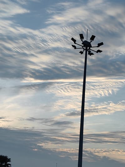 A tall, dark lamppost with multiple floodlights on top stands silhouetted against a dynamic sky filled with wispy, horizontal clouds. The sky transitions from a lighter blue at the top to a deeper gray-blue towards the bottom, with hints of warm light suggesting either sunrise or sunset. The clouds exhibit textured patterns, appearing elongated and streaky in some areas, and more layered and broken in others. At the very bottom left of the frame, the dark silhouette of a palm tree is partially visible. The overall scene is captured from a low angle, emphasizing the height of the lamppost and the vastness of the sky. The image appears to be taken outdoors in the city of Karnal, India.