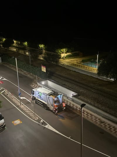 The image depicts an urban night scene in Pineda de Mar, Spain, captured from an elevated perspective. The primary subject is a white and green garbage truck positioned on an asphalt road, facing away from the viewer. The truck's rear compaction mechanism is illuminated, and two sanitation workers, dressed in orange and dark blue high-visibility uniforms, stand behind it on the road.

The road features white lane markings, a white arrow indicating a right turn, and a yellow "30" speed limit symbol painted directly on the asphalt. A white vehicle, possibly a van, is partially visible parked on the far left. To the right of the road, a pedestrian pathway with a distinct brown and white chevron pattern runs parallel to the street. A tall lamppost stands prominently in the foreground on the right.

Beyond the road and pedestrian path, two sets of train tracks traverse the scene horizontally on a raised embankment. An illuminated concrete underpass or tunnel provides access beneath these tracks. Dense vegetation, including bushes and trees, lines the areas bordering the tracks and road. Several additional streetlights illuminate the track area and the dark background. A blue circular sign with a white arrow and a rectangular yellow sign with red text are visible on poles near the tracks. The overall illumination is artificial, creating pronounced shadows.