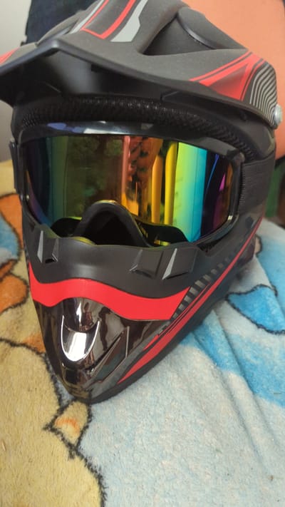 The image shows a close-up, eye-level view of a black motocross helmet with red and gray accents, resting on a patterned blanket. The helmet has a visor and goggles with a colorful, mirrored lens. The blanket is predominantly blue with yellow and orange patterns. The scene appears to be indoors, possibly in a home environment. There is no indication of the city of Zadebri, Ukraine.