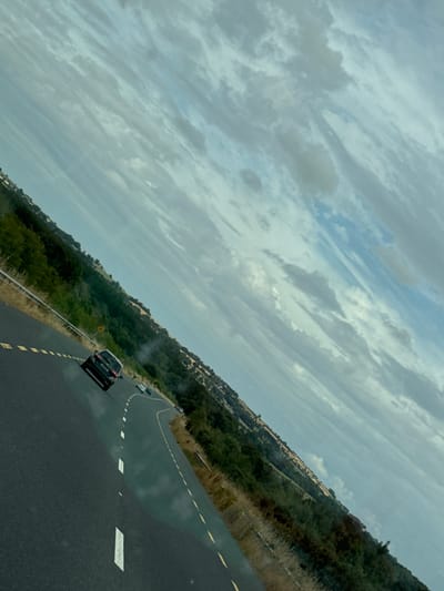 This is a tilted, medium-shot, outdoor, eye-level, horizontal, slightly hazy, natural light, day time photograph taken from a moving vehicle, showing a two-lane road in the countryside. The road is asphalt with a yellow dashed line on the right and a solid yellow line on the left. Several cars are visible on the road ahead. The landscape on both sides of the road consists of rolling hills covered in green grass and scattered trees. The sky is cloudy and appears to be overcast. The image is likely taken in the New Ross Municipal District, Ireland, given the characteristic landscape and vegetation.