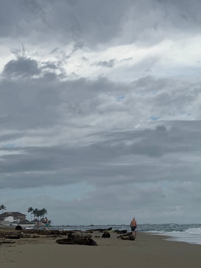 The photograph captures a beach scene in Cabarete, Dominican Republic under an overcast sky. The sky is dominated by thick, striated clouds. The beach is littered with driftwood and debris. In the foreground, a man stands near the water's edge and a family sits further down the beach. Palm trees and buildings are visible in the background, indicating the proximity of development to the beach. The waves are visible in the background, suggesting a rough sea.