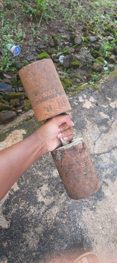 A person's hand is gripping the center of a homemade dumbbell. The dumbbell is constructed from two cylindrical metal weights, likely repurposed oil filters, rusted and marked with faded text. The weights are attached to a central bar, which is also visible as a white cylindrical object partially obscured by the hand. The background consists of a rough concrete surface with patches of moss and dirt, suggesting an outdoor environment. Scattered rocks and some green vegetation are visible in the upper portion of the frame. The scene appears to be located in Kemuning, Indonesia, based on the provided context. The lighting indicates it is daytime.