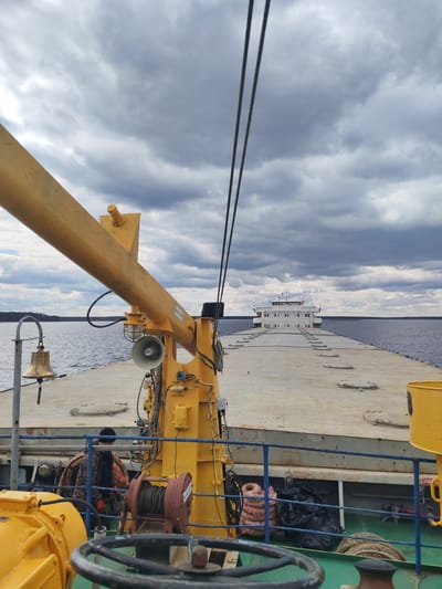 The image presents a working view from the deck of a vessel on a wide body of water, likely a river or large lake in the vicinity of Городище, Russia. The sky is heavily overcast with dark grey clouds, suggesting a cloudy day, but ample light indicates daytime.

In the foreground, a prominent bright yellow crane or boom arm extends from the left, fitted with a megaphone-like speaker, a light, and various electrical connections. A ship's bell hangs from a curved metal post on the far left. At the bottom center, a dark, weathered steering wheel is visible, while to its right, a red-brown winch with coiled rope suggests maritime operations. Blue railings separate the immediate foreground from the main deck. Behind these railings, partially obscured, a person wearing a dark beanie, a camouflage-patterned jacket, and orange gloves is seen, appearing to be working or observing. Stored below the deck level are pinkish coiled ropes and black garbage bags.

The main deck, which is wide and flat with several circular hatches, stretches towards the distant horizon. In the background, a large white multi-deck structure, resembling the bridge and superstructure of another vessel or a prominent section of a large barge, is clearly visible. The water extends to a distant landmass or treeline on both sides, reinforcing the inland waterway setting.

A white label with the text "P9350320" is visible on the yellow crane arm. A red sign, partially seen near the right edge on a yellow structure, appears to read "ПП". The overall scene captures a functional maritime environment, likely involving cargo transport or similar operations, under a characteristically cloudy sky.