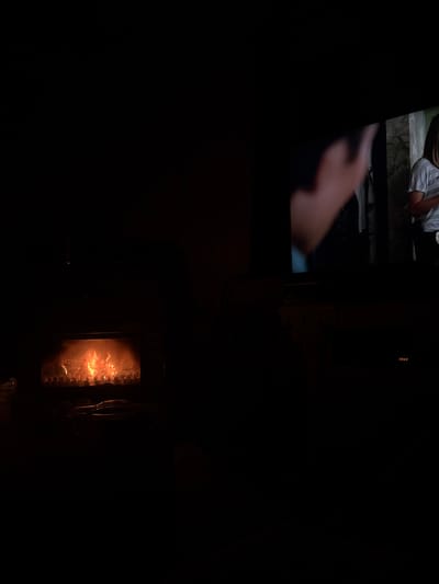 In a dimly lit space, the faint glow of a television screen displays an image of people outdoors. Below and to the right, a fireplace emits a warm, orange light, suggesting a cozy and intimate setting. The darkness envelops the scene, focusing attention on the contrasting sources of light.