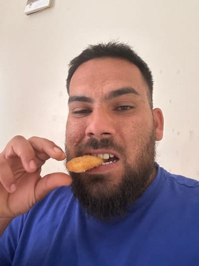 A man with a beard is eating a chicken nugget. He is wearing a blue t-shirt and is indoors. The wall behind him is white. There is a calendar hanging on the wall. It is difficult to determine the exact location in Boston, United Kingdom, based on the provided image.